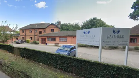 Google A red brick building and car park sit behind a green hedge. There is a parked car and a sign which says "Eastfield" in the foreground. 