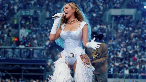 Beyonce is seen singing on stage during her Cowboy Carter tour in a silver and white outfit. She is wearing white fringed chaps. 