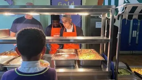 Sir Sadiq Khan is standing behind a glass hot food counter in a school as though he is serving food. He is standing alongside two staff members. He is looking down and appears to be talking. A boy has his back the camera and faces the food counter as though waiting to be served.