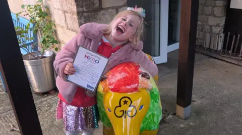 Submitted A six-year-old girl with blonde hair looks excited as she clings onto a painted lion sculpture and holds onto a "Girlguiding Hero" certificate. She is wearing white tights, a pink glittery skirt, a red zip-up hoodie and a pink fluffy coat.