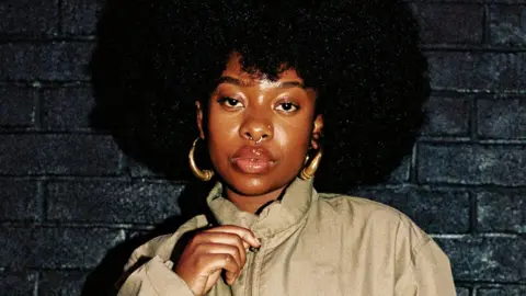Mariana Pires Meduulla with long black hair wearing gold hoop earrings and with her hand at the top of her zip on a beige coat. She is leaning on a black brick wall. 