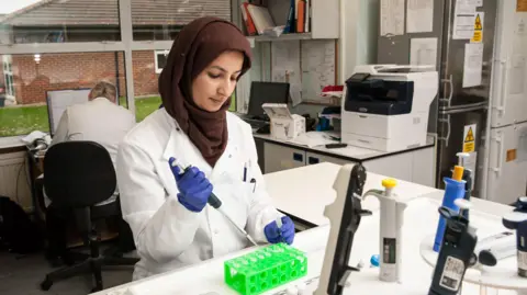 Epilepsy Society A laboratory setting where an individual in a white lab coat and gloves is carefully using a pipette to transfer liquid into a rack of small test tubes. The workspace is well-equipped with scientific instruments, including a printer, storage cabinets, and additional pipettes arranged on the bench. A large window behind the workstation lets in natural light and reveals a view of nearby buildings and greenery outside. In the background, another person is seated at a desk working on a computer.