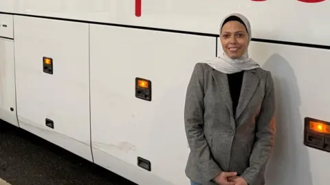 Fatima Benzbir wears a beige jacket, black top and white headscarf, as she leans casually against a coach.