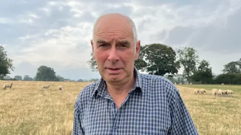 Photograph of Price, who is wearing a blue and white checked shirt, stood in a field with a serious expression. Behind him are about 10-15 white sheep. The field is covered in yellow-green grass, with trees lining the edge of the field. The sky is cloudy and overcast. 