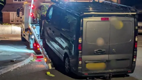 A grey van being hauled onto the back of a lorry on a dimly lit road.