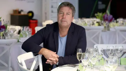 Getty Images John Torode sat at a dining table, wearing a suit. 