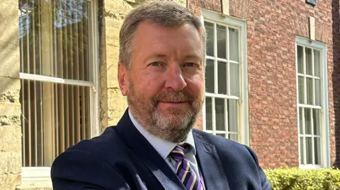 A man with blue eyes and short cropped grey-blond hair, with the same coloured beard and eyebrows. He is wearing a black, purple and cream striped tie from the collar of his blue shirt, over which is a navy blazer. He is standing in front of a red and stone-coloured brick building with Georgian-style windows.