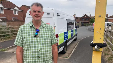 Mr Renshaw stood beneath the gate and next to the black bicycle lock that has been wrapped around the gate. A police van is parked behind him. He is wearing a green and white checked shirt with blue tinted sunglasses hanging from his shirt. 