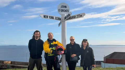 Lee Robinson Lee Robinson, wearing a yellow high-vis jacket and holding a red helmet, poses for a photo beside a white signpost at John O'Groats alongside three members of his land support crew—a young man and two women.