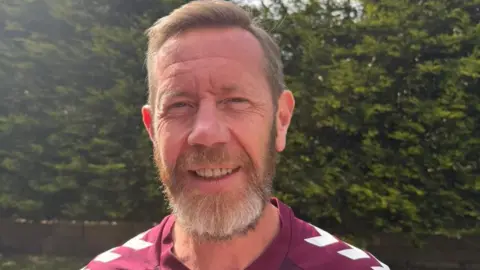 Colin Brown has short fair hair and smiles at the camera. He wears a maroon Hearts FC strip and is standing in a garden with a bush behind him