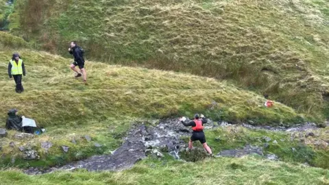 Fran Blackett  A runner is approaching a stream with a marshall in a high vis jacket watching on, above is another runner climbing the hill.