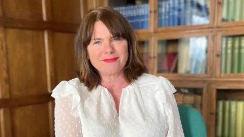 HMICFRS Michelle Skeer has brown shoulder-length hair, vivid pink lipstick and is wearing a white blouse. She is looking directly to camera and smiling slightly. There is a row of wooden bookshelves behind her.