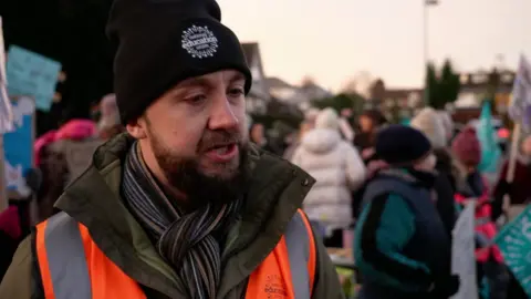 A man with a beanie hat stands on the picket line outside a school. He is wearing a green coat, scarf and hi-vis jacket.