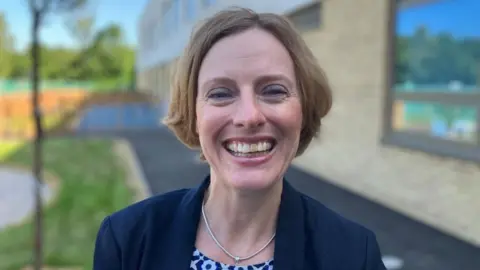 Sam Read/BBC Catherine Assink standing outside a school building, smiling at the camera, wearing a silver necklace, blue jacket and patterned top. She has mid length fair hair.