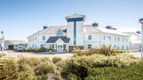 Hospice Isle of Man The Hospice building is cream and wraps around in a hexagon shape so you can see the three front sides. There are shrubs and bushes in the forground and it is a sunny day. 