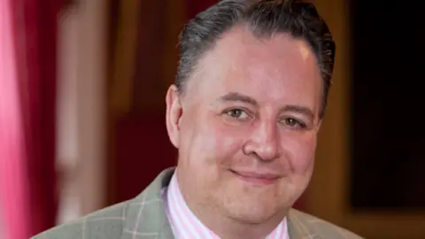 Steven Moore is wearing a green chequered suit with a pink shirt. He is smiling at the camera and has dark hair. 