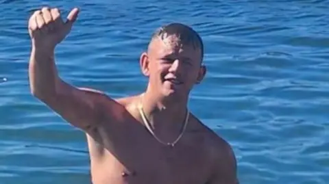 Lewis Smith stands in the sea bare-chested. He has his right arm raised with a thumbs up and is smiling. He has a gold chain around his neck. 