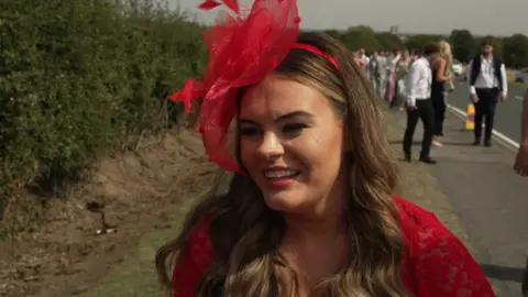 Jodie Crawford, with long brain hair, is wearing a red fascinator and a red dress. Behind her is a long queues of fellow racegoers.