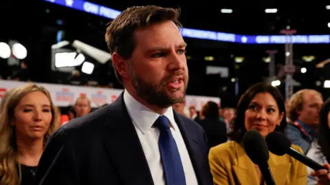 Reuters A white man with a beard and navy suit stands in front of a couple of hand-held microphones while two women look on.