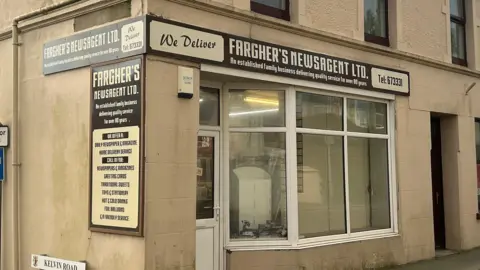 The exterior of the shop, it has brown and cream livery which reads "we deliver" and states the name Fargher's Newsagent Ltd.