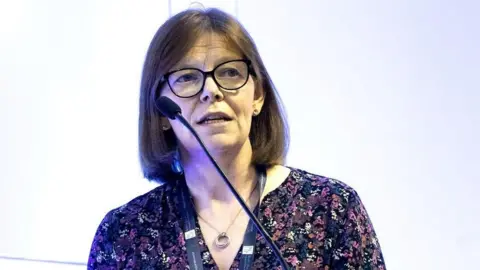 RGCP A close-up image of Dr Ursula Mason speaking at a podium with a large mic. She has shoulder-length brown hair and is speaking into a microphone. She is wearing a pair of dark, circular glasses, a patterned shirt and she is standing in front of a light wall looking out into the crowd.