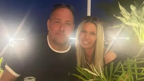 Family handout/GoFundMe A man wearing a T-shirt and sitting next to a woman with blonde hair and wearing a black top. They are both smiling at the camera.