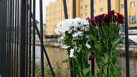 Two bunches of flowers, red and white, tied to a black bridge railing with water and apartments visible in background