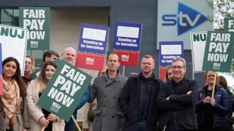 A group of people wearing coats holding big green and blue National Union of Journalists (NUJ) signs at a picket line with the STV building behind. 