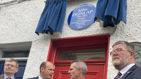 Shows a blue plaque with the words John Dunlap and four men standing below it