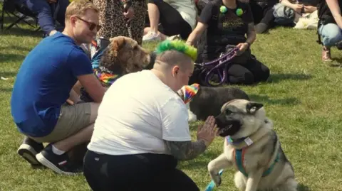 BBC/John Bray Pride festival-goers with their dogs in the gardens in Leamington, showing one dog in a rainbow collar with their foot up to their owner