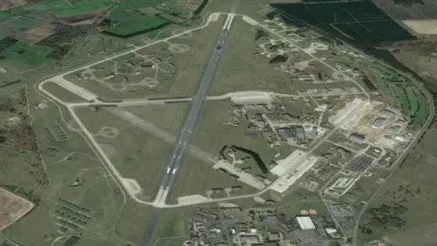 Google Earth Aerial view of an air base. Runways, hangars and buildings are visible. There are also hardened shelters for fighter aircraft.