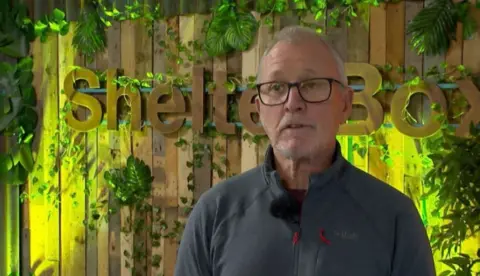 A man with short grey hair, glasses and a blue zipped-up top stands in front of a wooden, illuminated Shelterbox logo surrounded by plants.