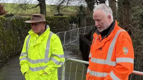 Supplied Nick Hampson and Malcolm Perrin stand side by side on a narrow grey path bordered by a stone wall on one side and a black metal fence on the other. A temporary grey metal fence has been erected down the middle of the walkway. Hampson, who is wearing a brown hat and a yellow high-vis jacket, is looking off into the distance. Perrin, who has short grey hair and a beard, is also wearing an orange high-vis jacket and is looking in the same direction.