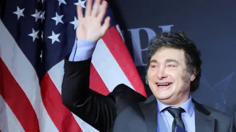 Argentina's President Javier Milei waves in front of an American flag, wearing a dark blue suit.