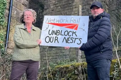 Supplied A woman in a green sweater and a man in a blue coat and cap hold up a sign near the viaduct which reads NETWORK RAIL UNBLOCK OUR NESTS