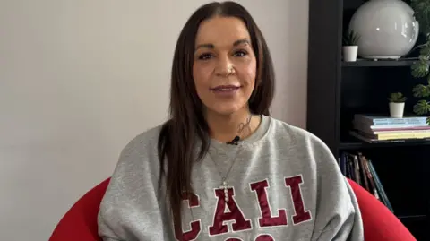 A woman from Gloucester who previously used ketamine warns about ‘k‑cramps’ and long‑term bladder damage. She has long dark hair and is smiling. She is sitting in a red armchair with a grey wall and black bookcase behind her. She is wearing a grey sweat top with Cali on it in red letters and a diamante cross necklace.