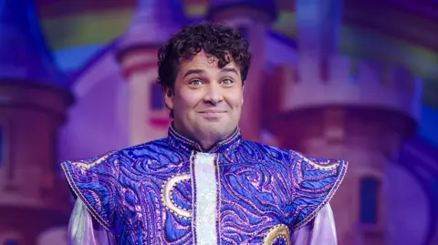 Joe McElderry is on stage and smiling sheepishly. He has short, curly, dark hair. He is wearing a costume which is gold and purple and has pointy shoulders.