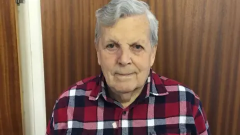 John Lloyd John Lloyd, wearing a red, black and white plaid shirt. He has grey hair and is looking towards the camera