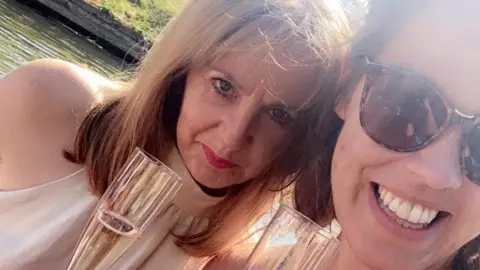 Ellishia Lauder Two woman are posing for a selfie. They are holding Champagne flutes and are outdoors. One of the women is wearing glasses, the other is wearing a light-coloured dress.