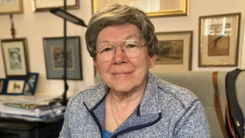 A woman wearing a grey fleece and glasses, smiling in her living room with photos on the cream wall behind her