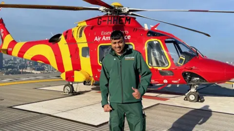 University of Hertfordshire Jaabir Booly, wearing a green uniform, with top and bottoms, holding his left thumb up, standing in front of a red helicopter, with yellow stripes on it. 
