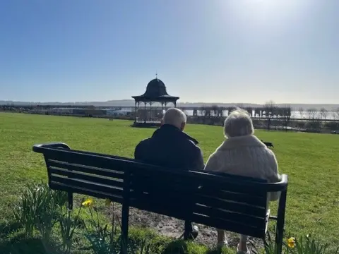 Elaine Morrison A wide grassy park on a sunny day, with two people sitting on a black bench facing away from the camera. In the distance stands an ornate bandstand and a large body of water. Daffodils bloom near the bench, and the sky is clear and bright.