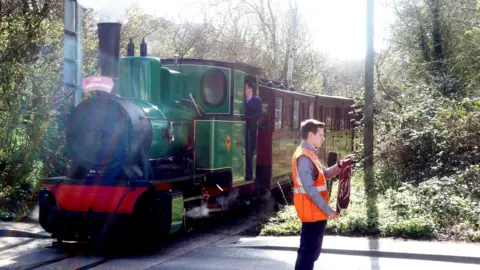 David Barrow A man standing in the road, side on, holding a flag, wearing an orange high-vis jacket. A train is passing by, with a person in the carriage. Foliage is either side of the train. 