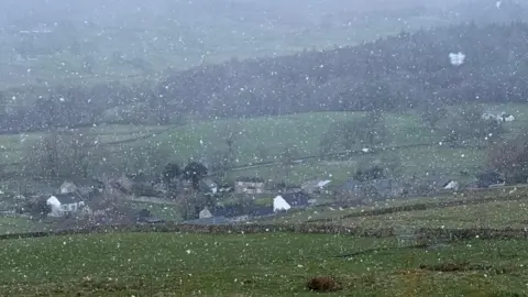 Toby the Gardner Lots of flecks of snow falling all around the photo. There is a valley in the centre of the photo with a collection of detached houses. Behind them is a stretch of fields with a woodland covering the centre.