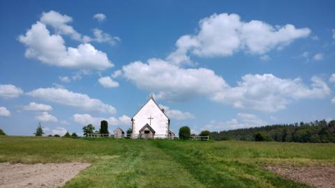 THURSDAY - The church at Idsworth - a small white chapel with a cross on the apex wall - photographed from a distance with green  grass surrounding it and sunny blue skies above with fluffy white clouds
