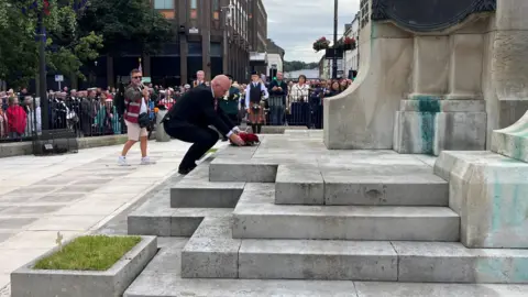 An older man wearing a black suit is bent over in front of a large stone war memorial. He is placing a poppy wreath on one the memorial's steps. There's a large crowd in the background watching him. 