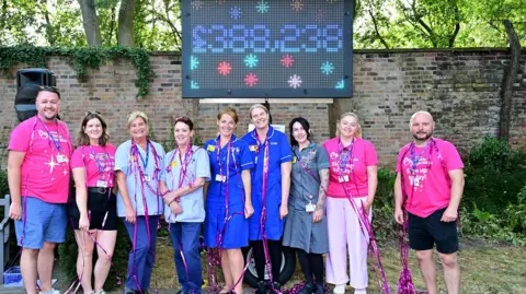 Nick Rhodes A group of hospice staff stood in a garden in front of an electronic totaliser showing the figure £388,238