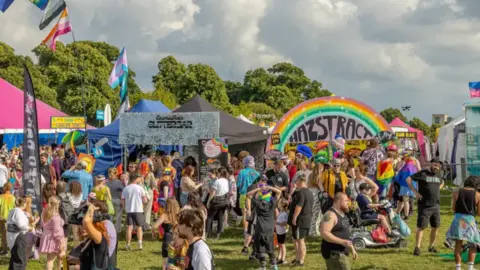 Bristol Pride / Dan Weill Photography A crowd of people wearing colourful outfits standing on grass with colourful stalls and flags in the background. 