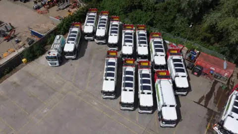 Dozens of white waste and recycling lorries parked on a large expanse of tarmac. Solar mats can be seen on the roof of each vehicle.