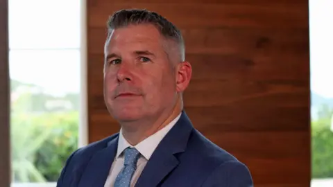Headshot of David Cattie. He has short dark hair and is wearing a blue suit, white shirt and blue tie with white polka dots. He is clean-shaven. He is pictured indoors, in front of a wooden panel.